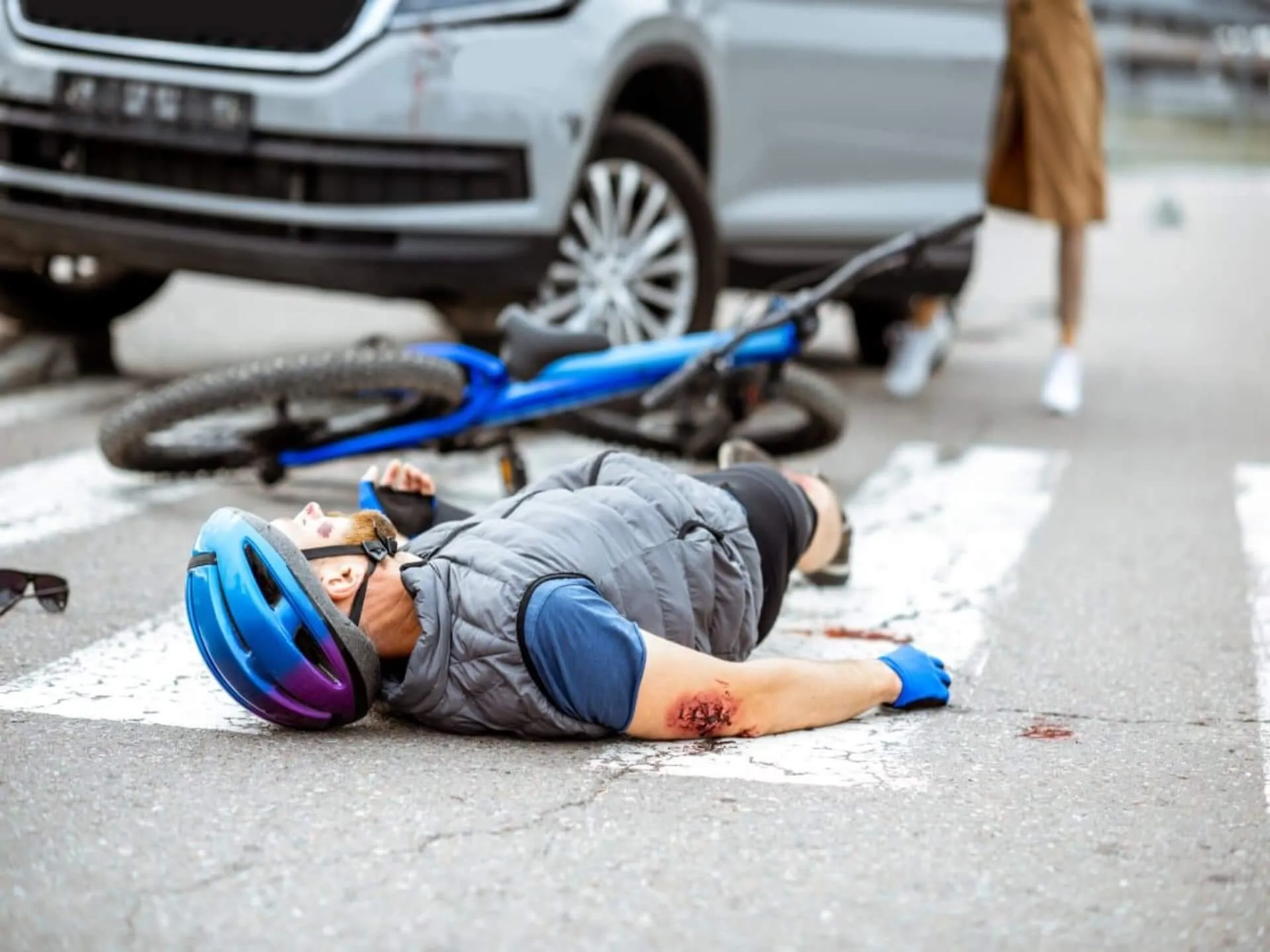 A helmeted cyclist lying beside a fallen bicycle near a car.