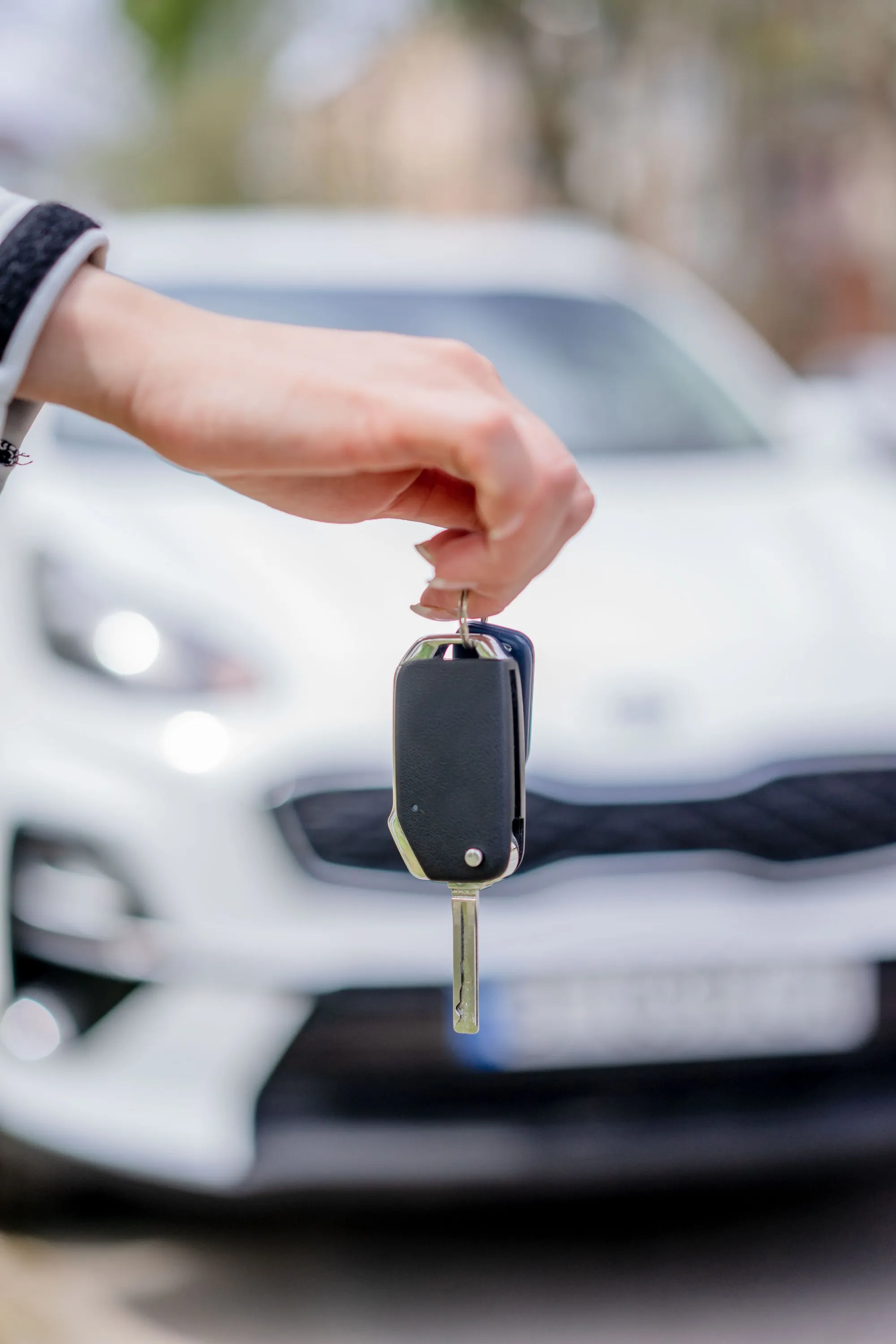 A hand holding a car key in front of an out-of-focus white car.