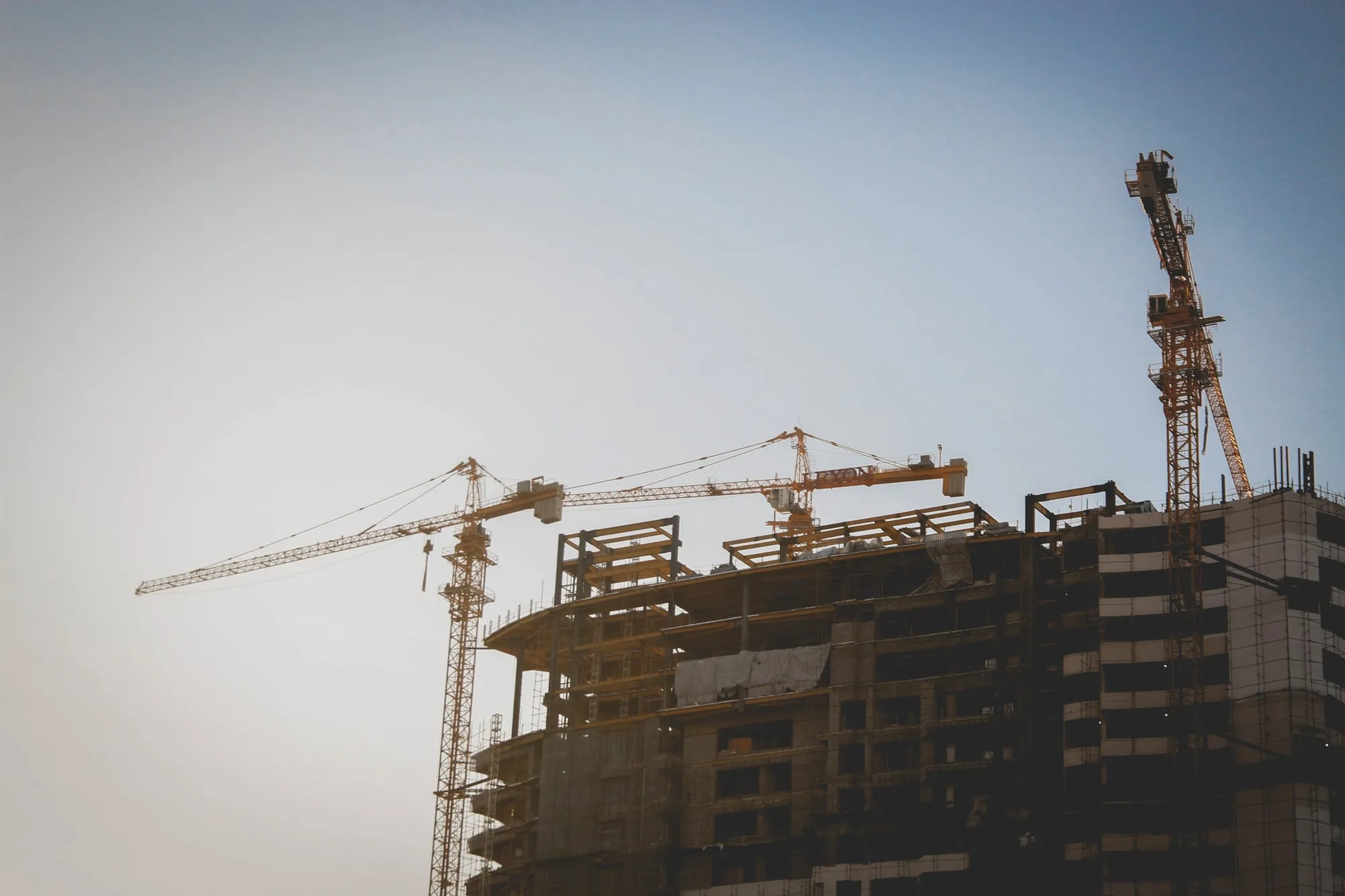 Cranes above a building under construction against a pale sky.