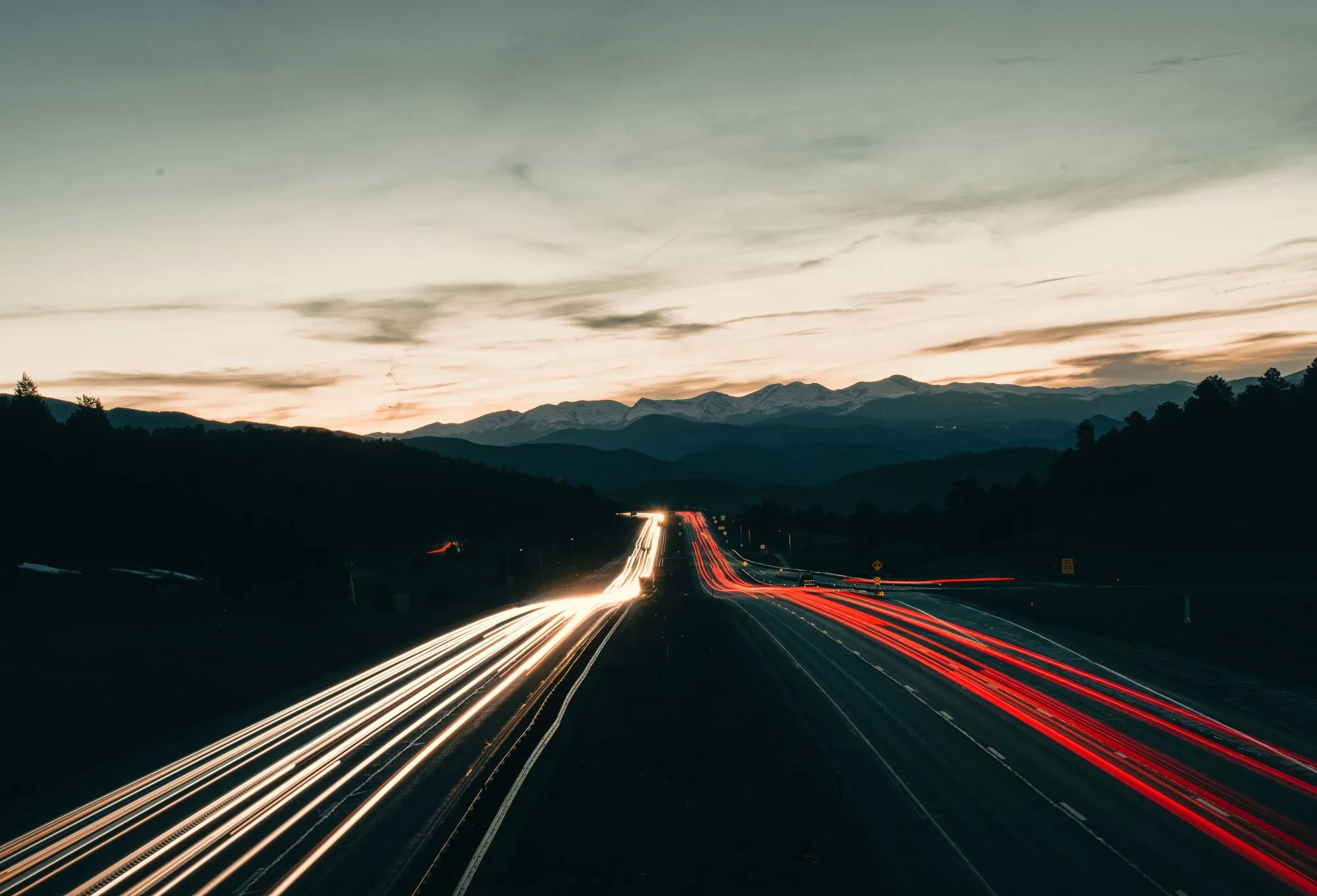 Light trails from traffic moving in both directions on a divided highway at dusk, with mountains in the distance.