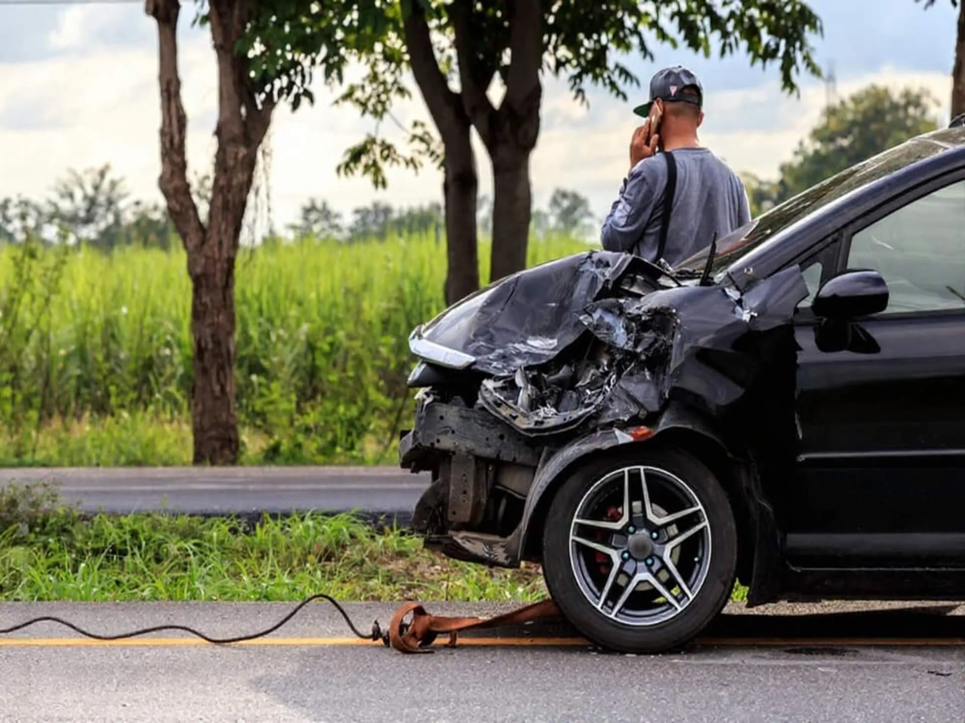 A damaged black car with a person using a phone beside the road.