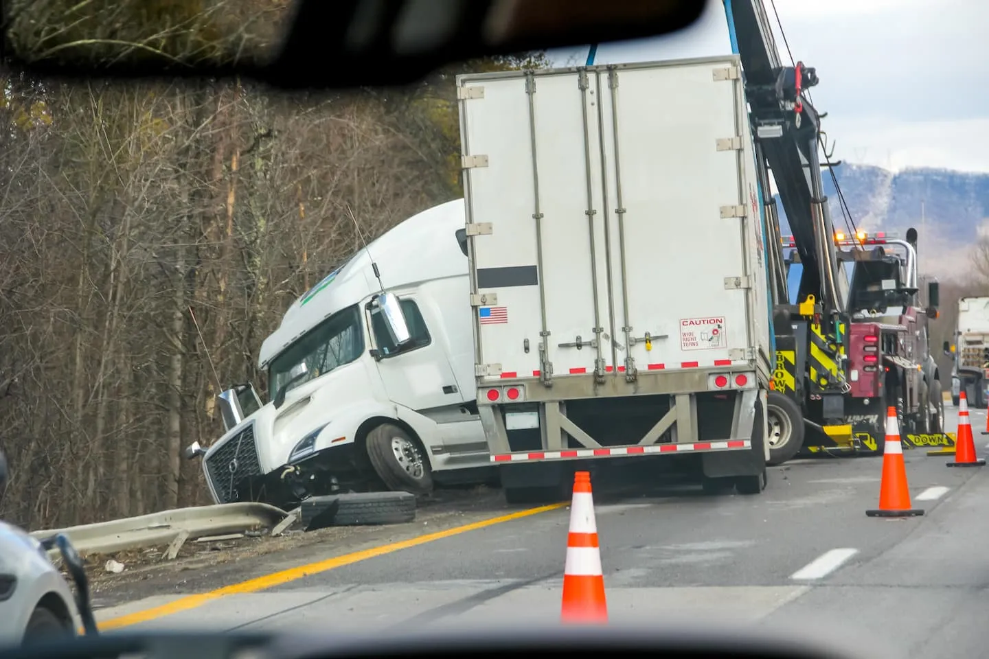 A white 18-wheeler with its cab off the highway beside a damaged guardrail, as a heavy-duty tow truck and traffic cones mark the crash scene.