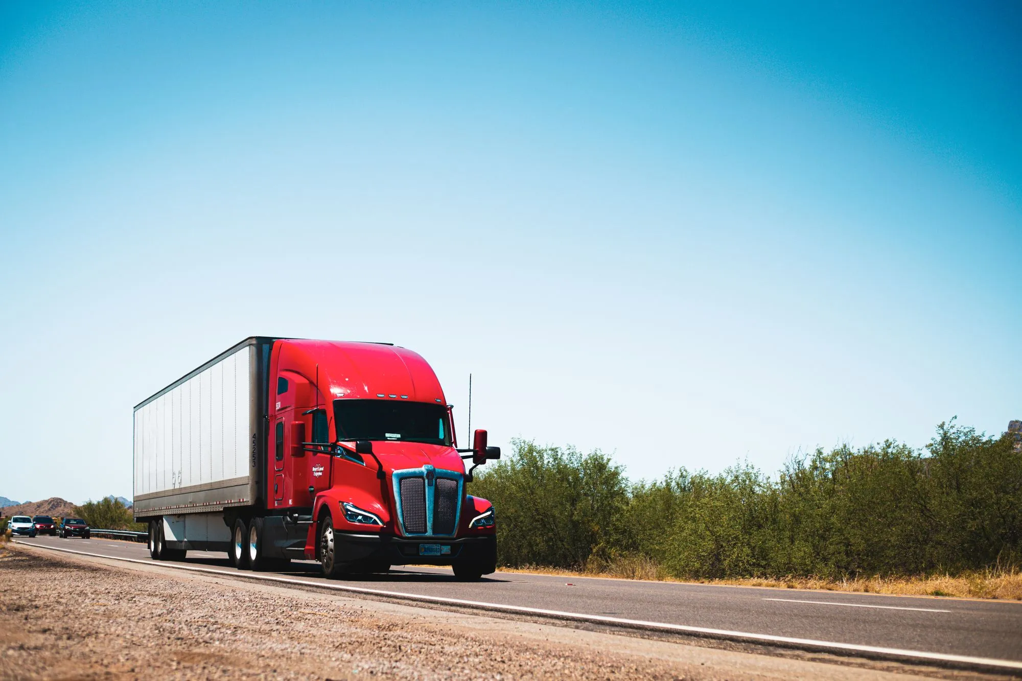 A red semi-truck pulling a plain white trailer on a two-lane highway through dry brush.