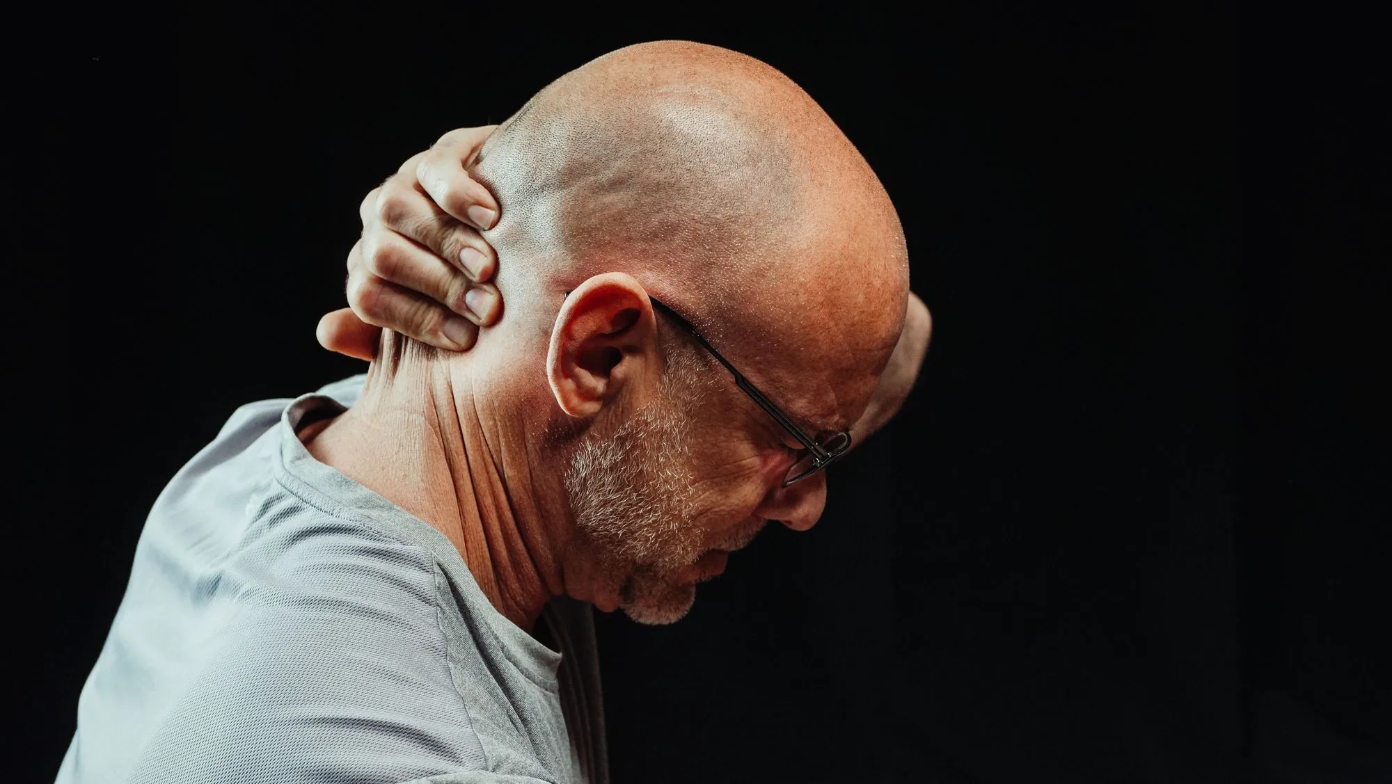 A man with his head bowed holds the back of his neck against a dark background.