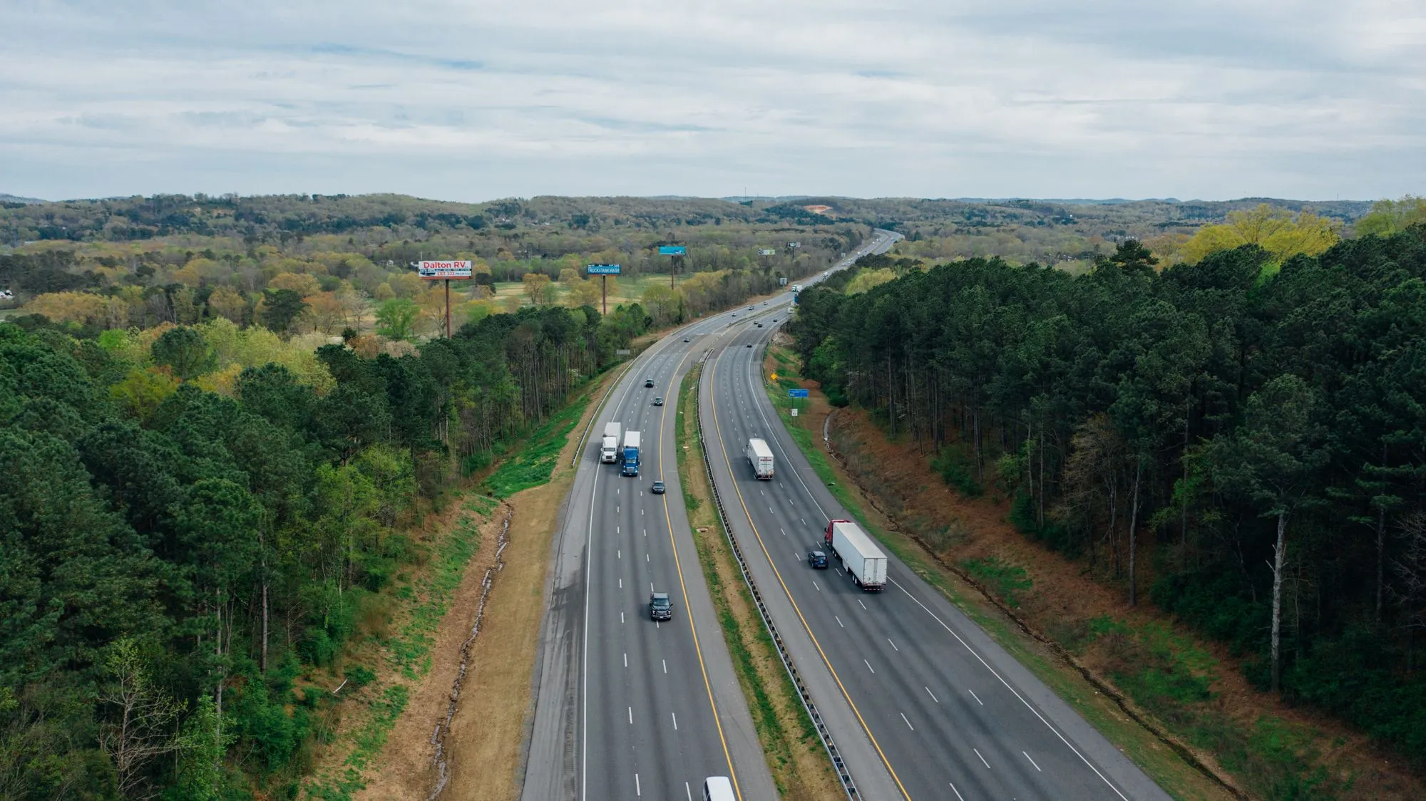 Aerial view of a four-lane interstate through pine forest, with semi-trucks and cars traveling in both directions.