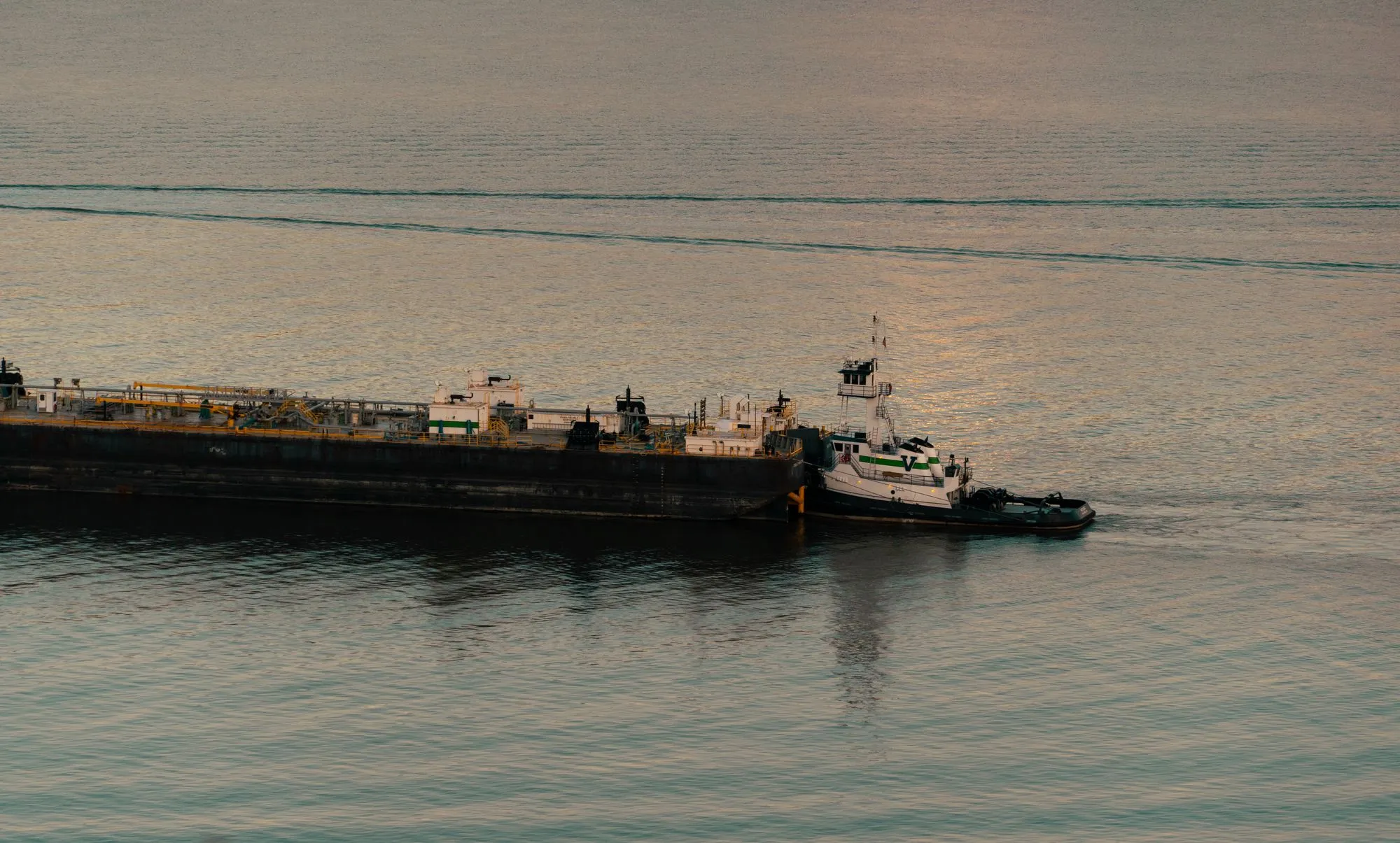 A tugboat moving an industrial barge across calm water at dusk.