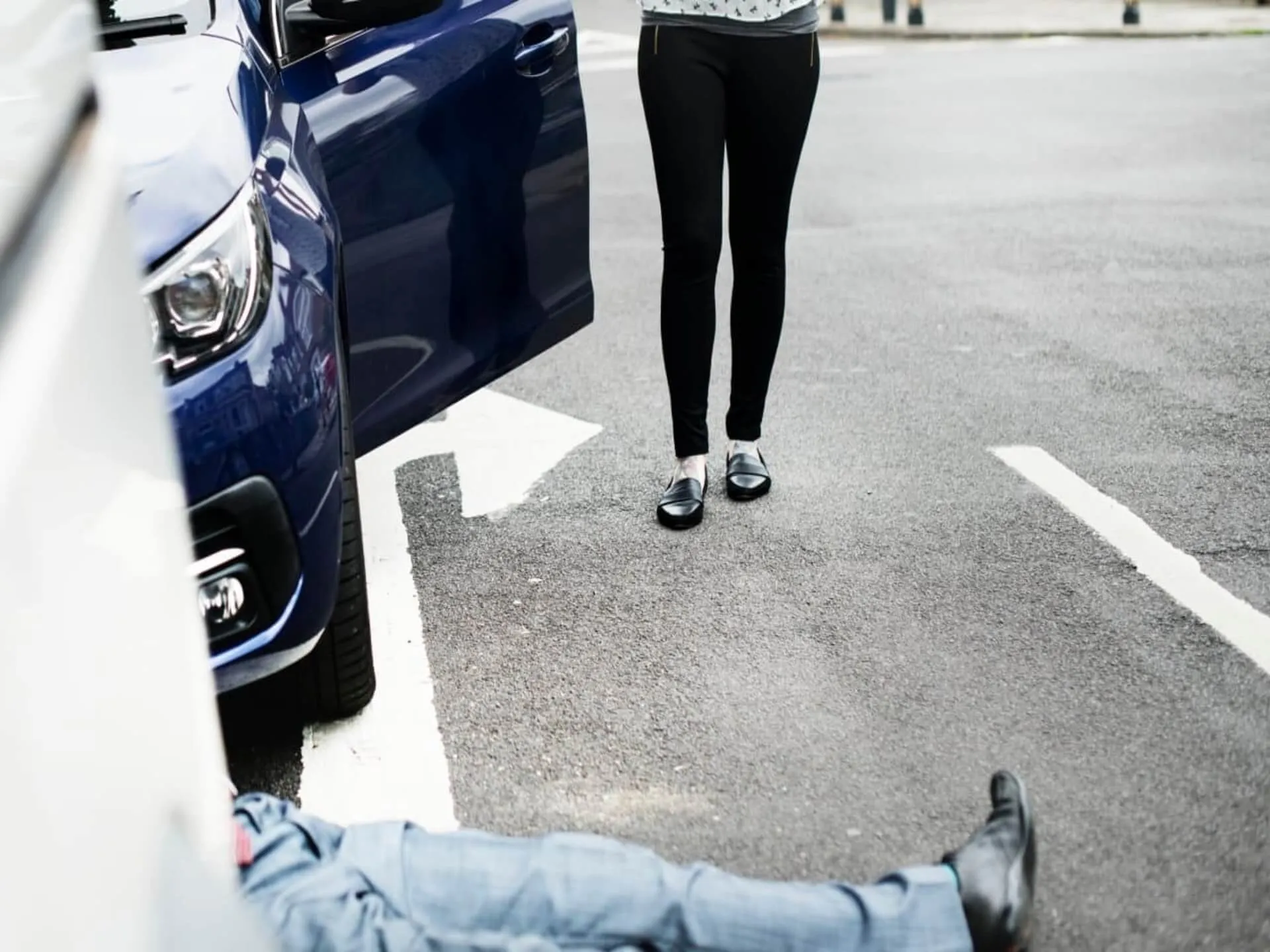 A person lying on the roadway beside a car, with another person standing nearby.